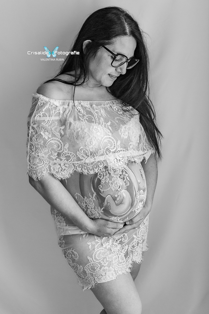 Mamma incinta in abito bianco di pizzo, tre quarti, sorriso verso il pancione, occhiali, gonna corta e belly painting che si intravede, gamba piegata su sfondo bianco