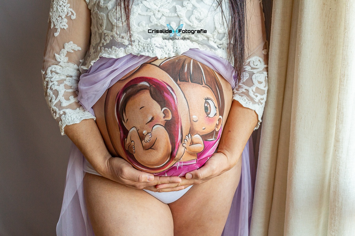 Primo piano sul pancione di una mamma con belly painting realizzato a mano da un'artista. Mamma con abito bianco in pizzo, manica tre quarti, gonna in tulle viola aperta sulla pancia. Entrambe le mani sotto il ventre, dita intrecciate. Luce morbida laterale proveniente dalla finestra, tenda sullo sfondo, atmosfera intima e delicata