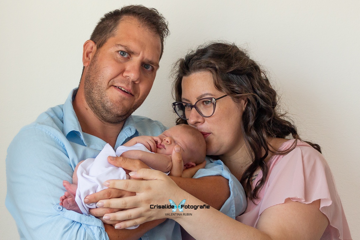 Ritratto familiare con papà con camicia azzurra e barba curata, mamma con abito chiffon rosa e occhiali che bacia la testina della neonata avvolta in tessuto bianco, sfondo bianco - fotografia newborn professionale a domicilio