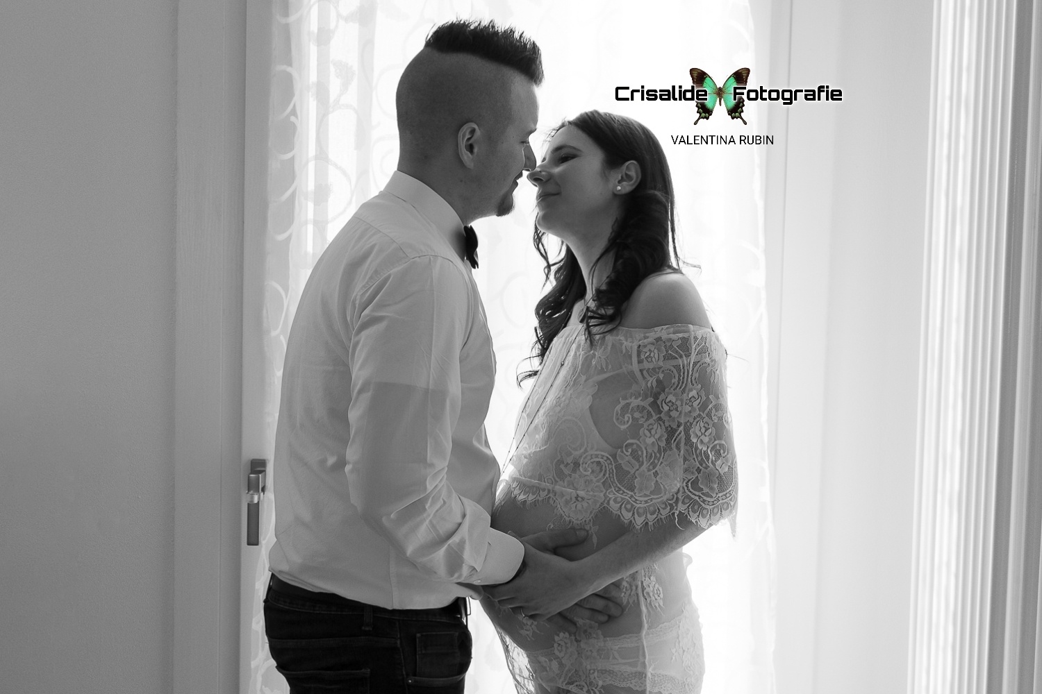 Coppia in bianco e nero che si bacia tenendosi per mano, lui con camicia bianca e papillon nero, lei con abito in pizzo bianco, luce naturale da una finestra con tenda