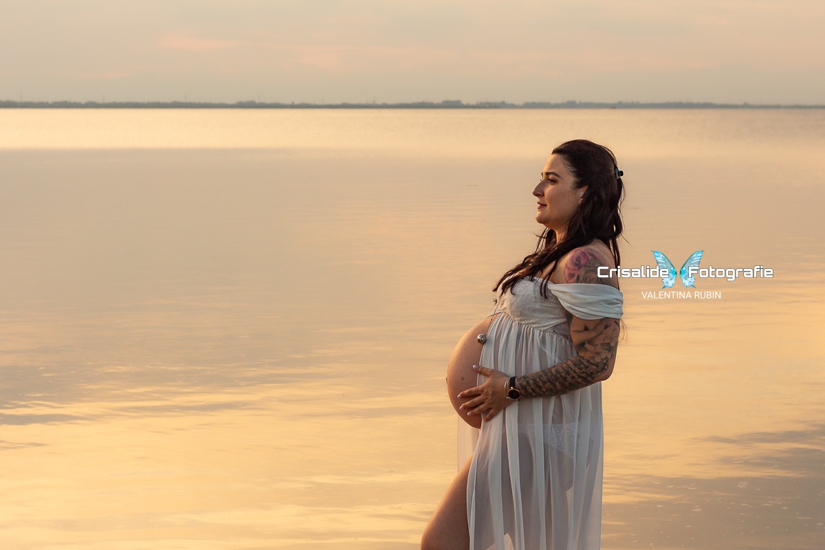Mamma incinta di profilo in abito di chiffon bianco aperto sul pancione, con il mare sullo sfondo e riflessi rosa pastello sull'acqua