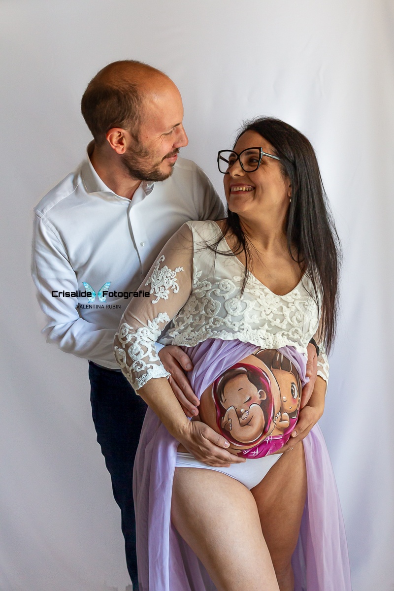 Papà dietro sulla sinistra e mamma davanti sulla destra, si guardano sorridendo. Entrambe le coppie di mani sul lato del pancione. Papà con camicia bianca e pantaloni blu scuro, leggermente pelato con barba corta. Mamma con abito bianco in pizzo, manica tre quarti, gonna in tulle viola aperta sulla pancia e belly painting colorato. Capelli lunghi lisci neri e occhiali. Sfondo bianco, atmosfera intima e gioiosa di famiglia