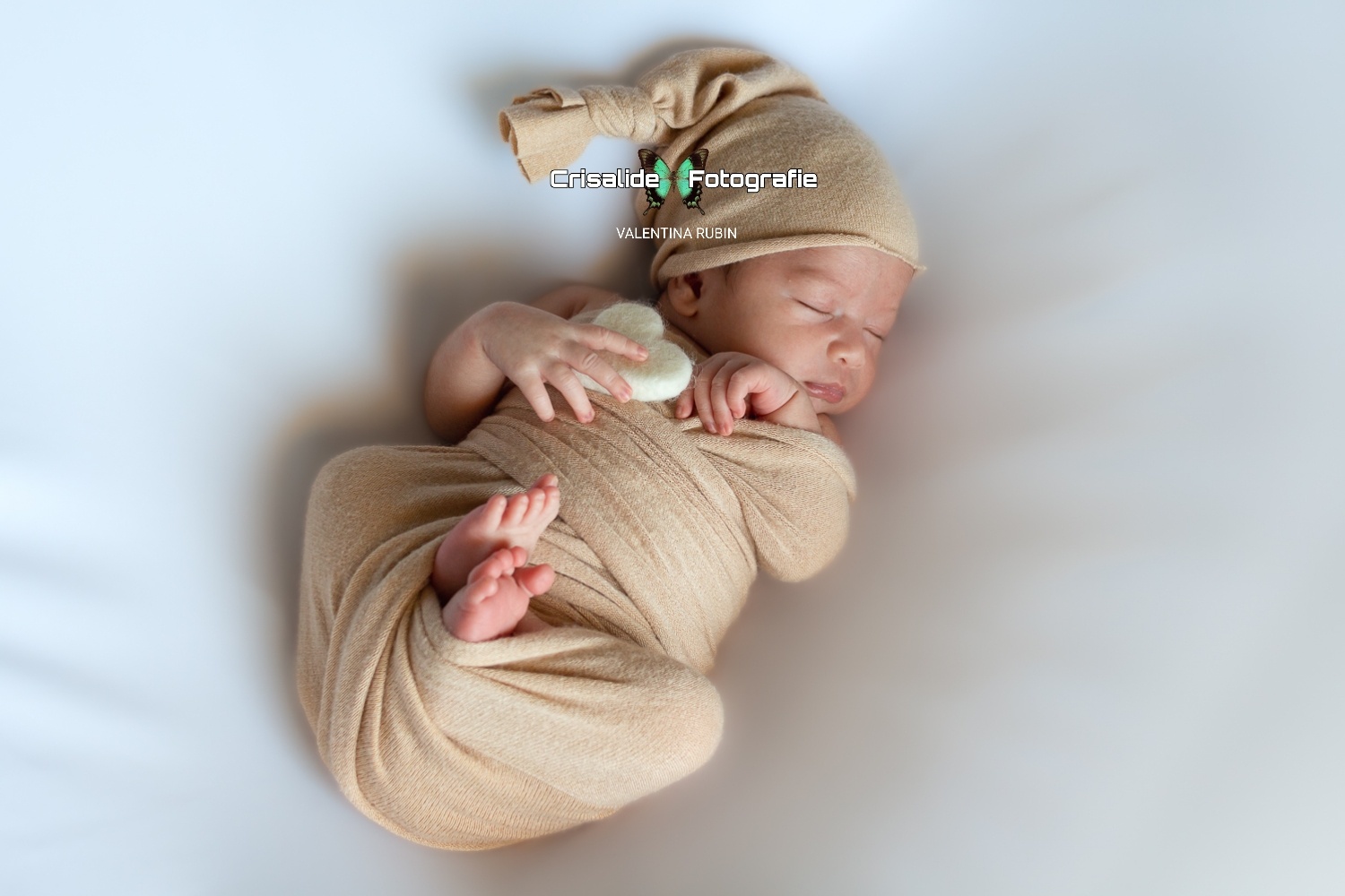 Neonato che dorme supino avvolto in fascia di cotone beige, con gambine rannicchiate, manina sulla guancia, cuore bianco in feltro nella mano, berrettino coordinato e sfondo bianco - fotografia newborn professionale a domicilio