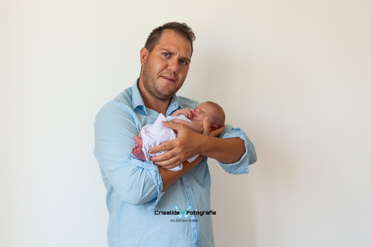 Papà con camicia azzurra che tiene in braccio la neonata con fascia bianca, mentre sorride all'obiettivo; neonata dorme su sfondo bianco