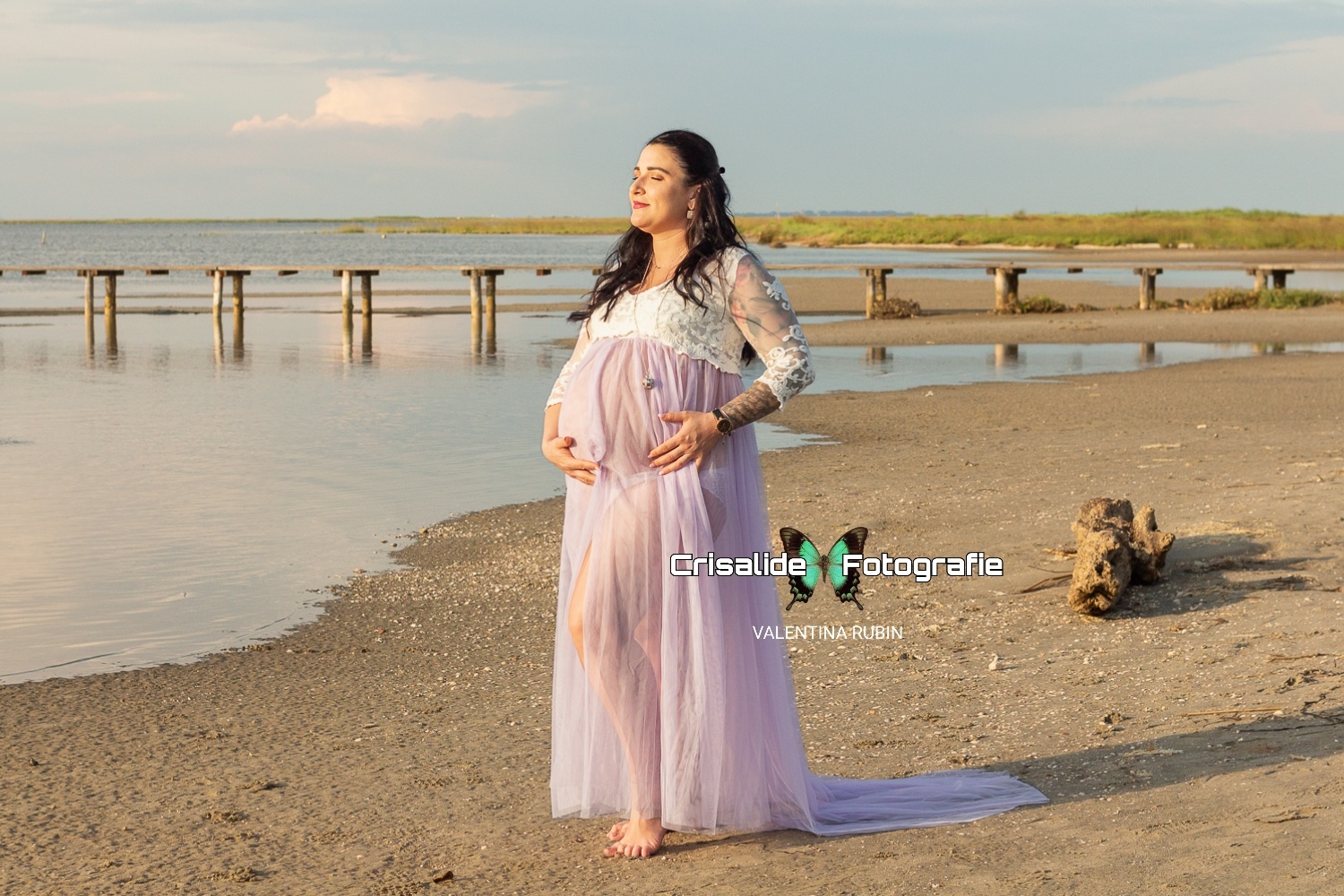 Mamma incinta con abito bianco in pizzo e gonna in tulle violetto, fotografata di tre quarti mentre tiene le mani sulla pancia. Ha gli occhi chiusi rivolti verso il sole. Sfondo con mare a sinistra, spiaggia con un tronco a destra, pontile di legno in lontananza ed erba. Cielo azzurro con una nuvola bianca