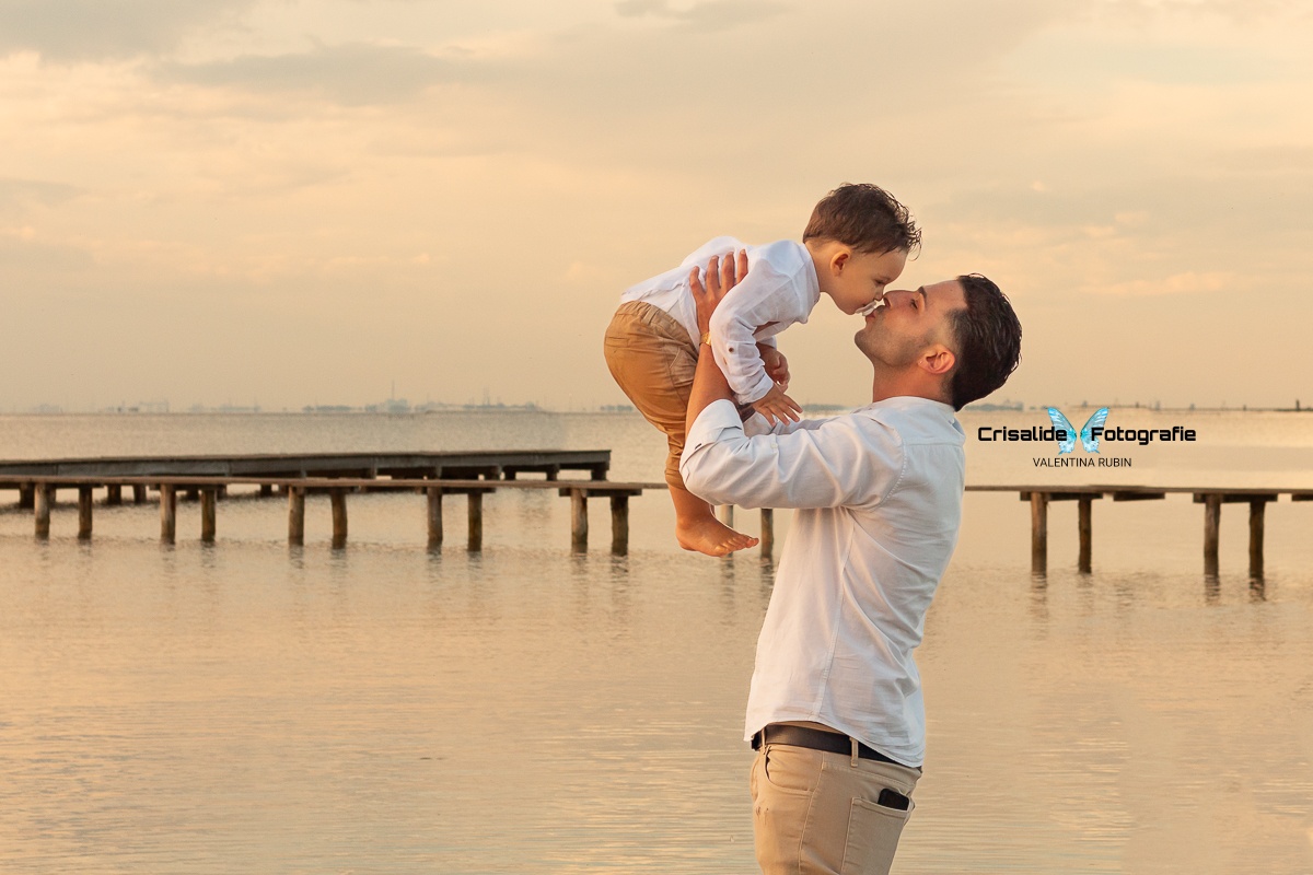 Papà in camicia bianca e pantaloni beige tiene in braccio il bambino vestito uguale, mentre lo bacia, sfondo pontile e mare con nuvole
