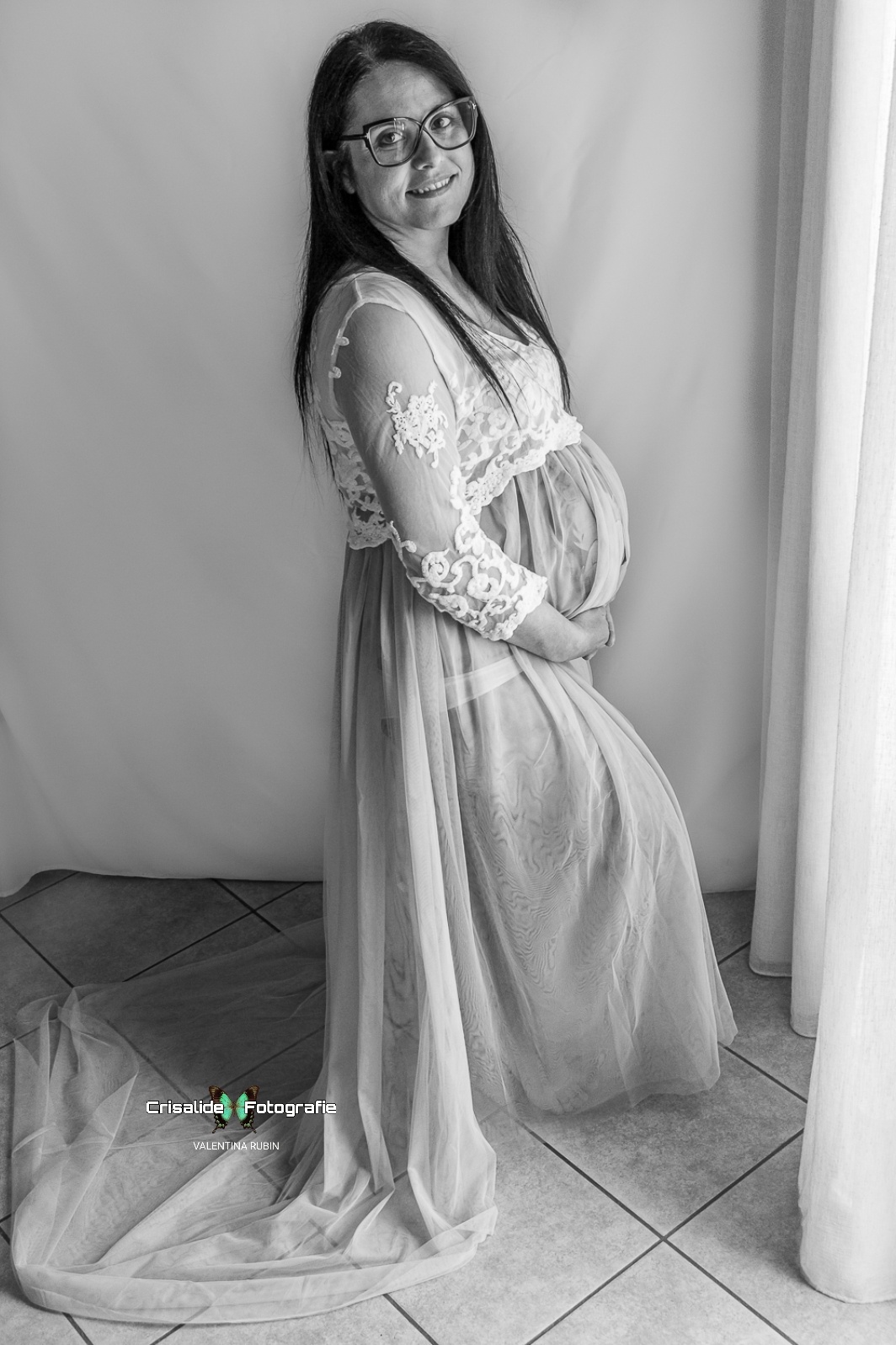 Mamma incinta di profilo in abito bianco di pizzo e gonna lilla in tulle, sguardo in camera e sorriso, occhiale, luce dalla finestra sulla tenda