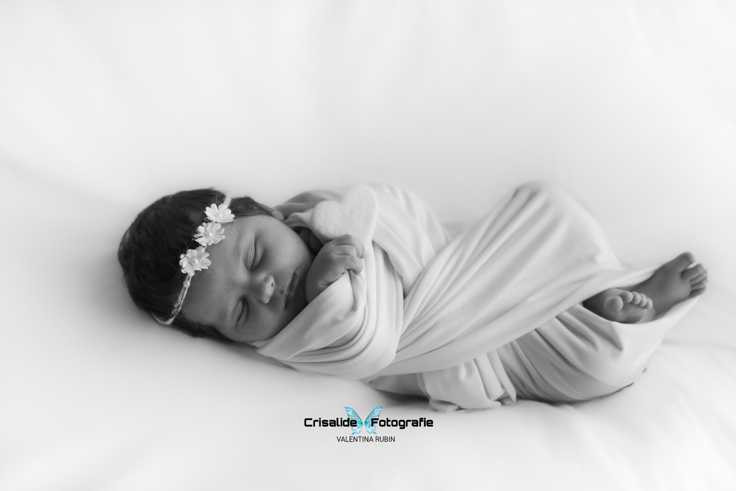 Neonata che dorme fasciata di bianco, a figura intera, con cuore bianco in mano e coroncina di fiori, scatto in bianco e nero