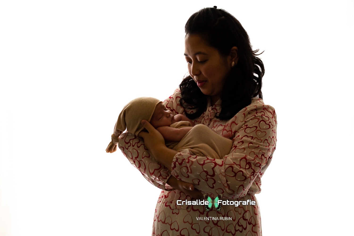 Mamma in abito beige con cuori rossi tiene in braccio il neonato fasciato in beige con berretto coordinato, sfondo bianco