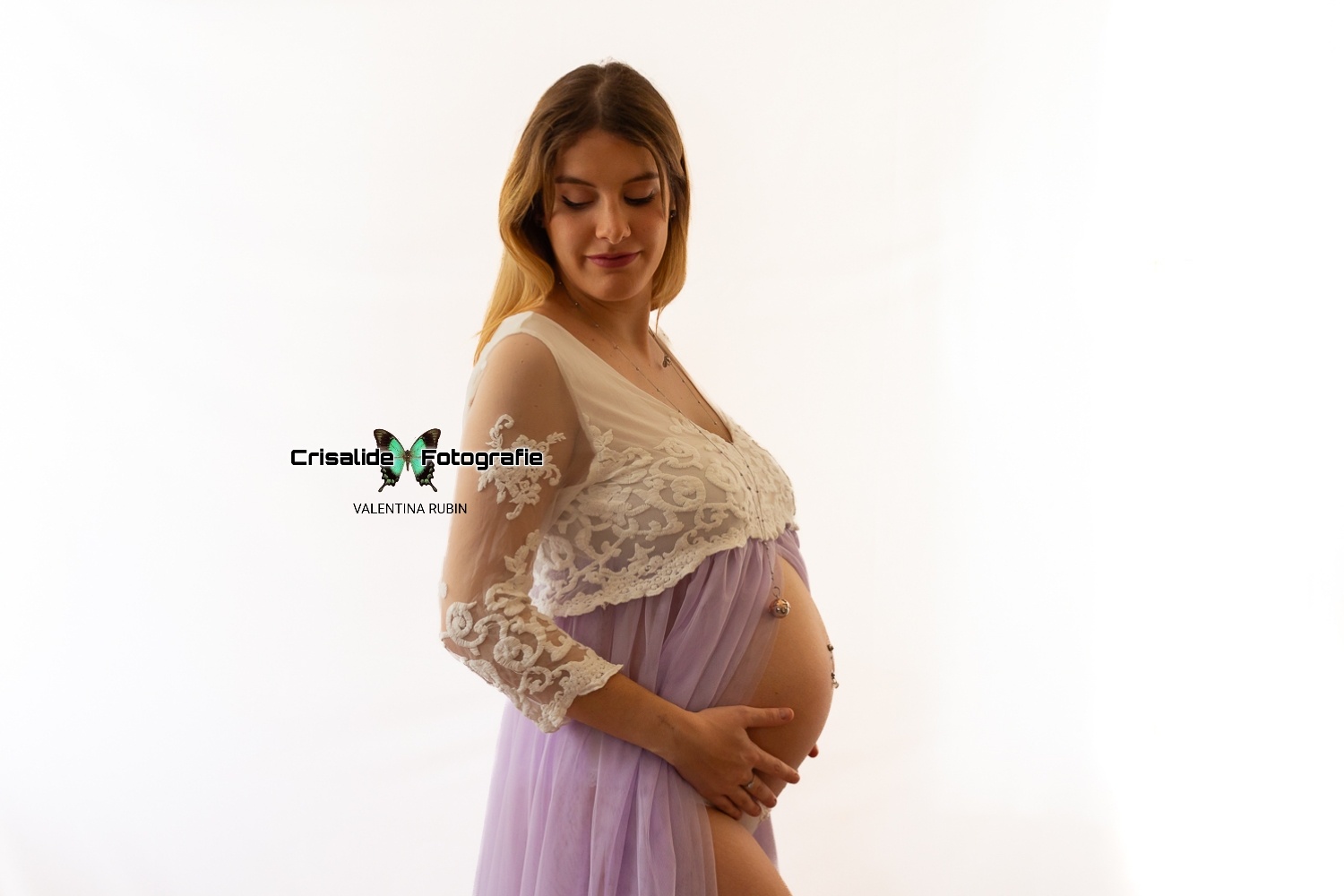 Donna incinta di profilo con vestito bianco e gonna in tulle violetto, che accarezza la pancia e sorride davanti a sfondo bianco