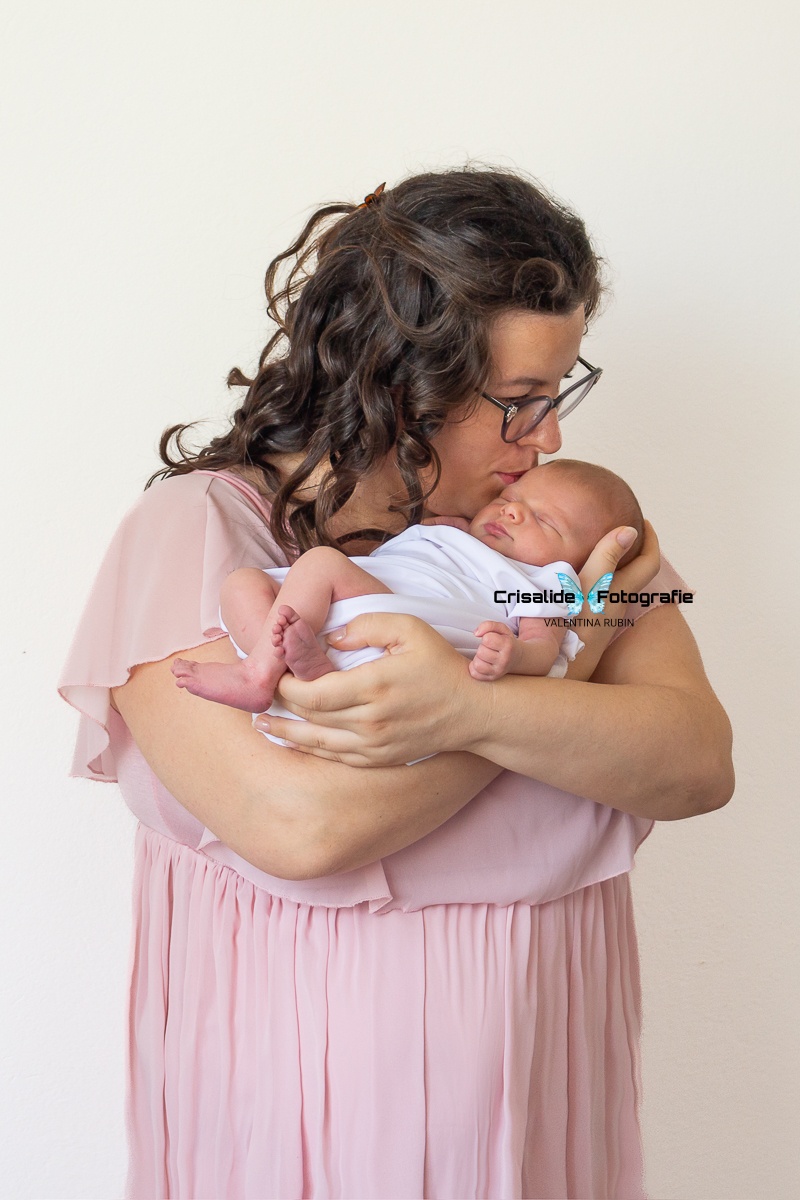 Mamma con abito rosa in chiffon tiene in braccio la neonata fasciata di bianco mentre dorme, la bacia sulla testina. La mamma ha capelli castani ondulati e occhiali. Sfondo bianco. Servizio fotografico neonati a domicilio
