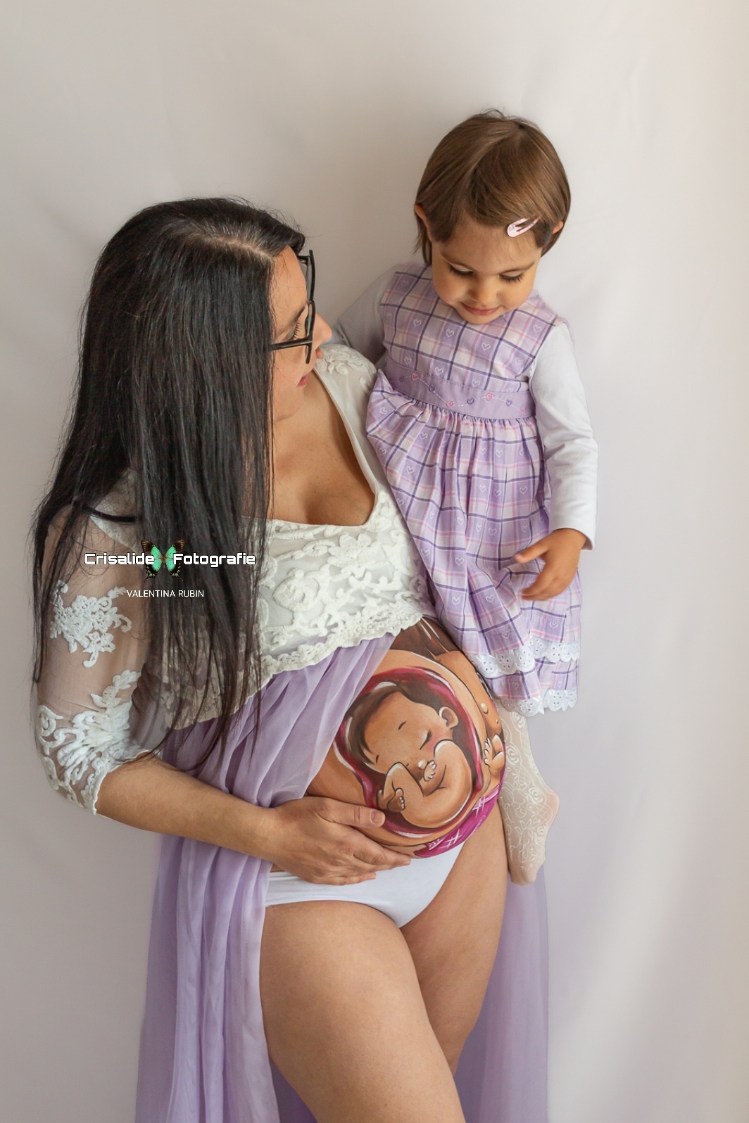 Mamma incinta con abito bianco in pizzo e gonna in tulle violetto, aperta sulla pancia, tiene in braccio la figlia di 2 anni vestita a quadretti viola e manica lunga bianca. Belly painting colorato sul pancione. La mamma con capelli neri lunghi lisci e occhiali guarda la bambina, mentre la bambina guarda il pancione. Mano della mamma sotto la pancia. Sfondo bianco, atmosfera gioiosa e familiare