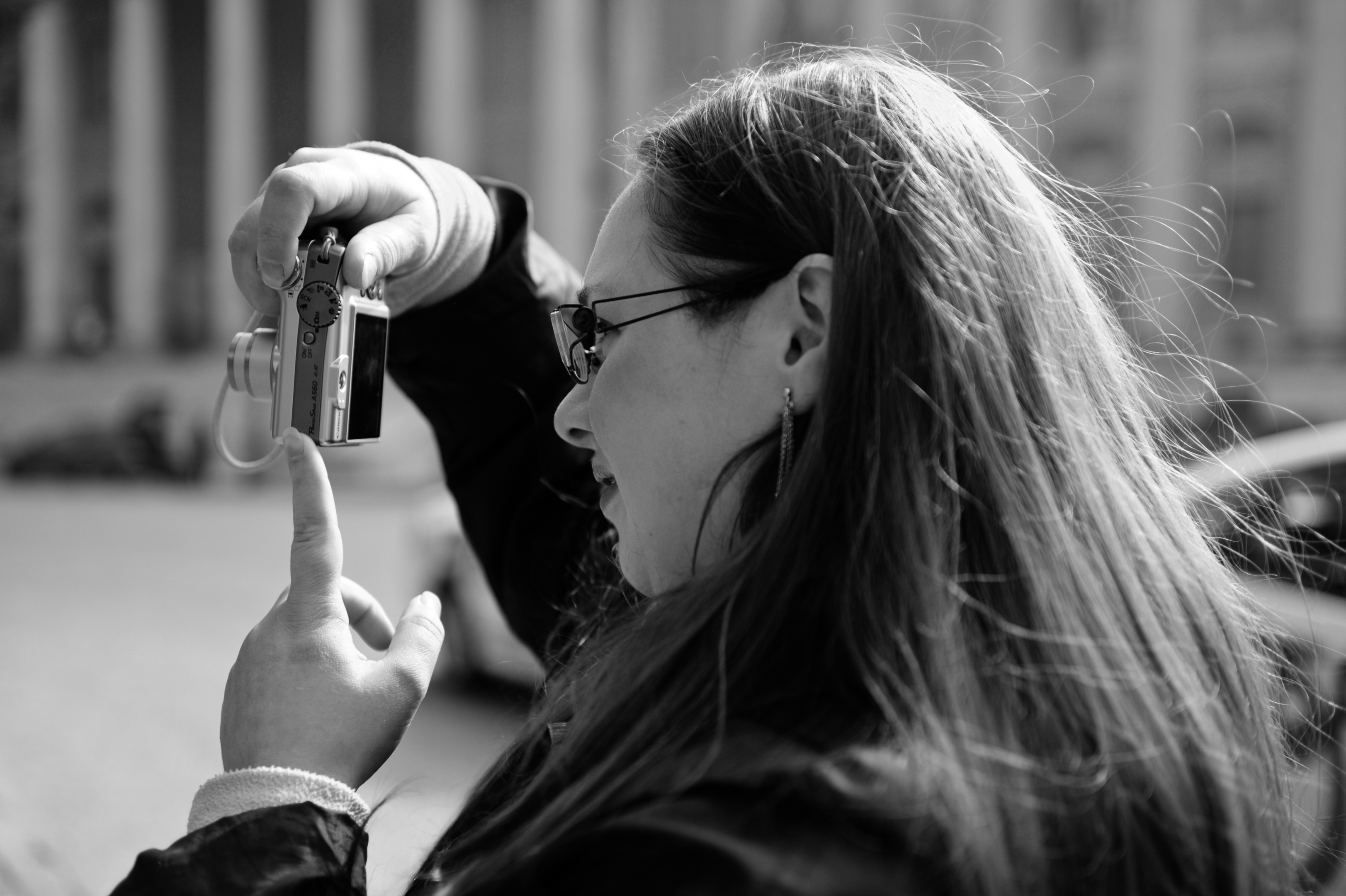 Valentina Rubin di profilo, in bianco e nero, capelli lunghi sciolti, occhiali e orecchini, mentre tiene con la mano destra una macchina fotografica compatta verticale e con la sinistra la sostiene;  luce retroilluminata illumina capelli e mani; sfondo sfocato con colonne di un edificio a Verona