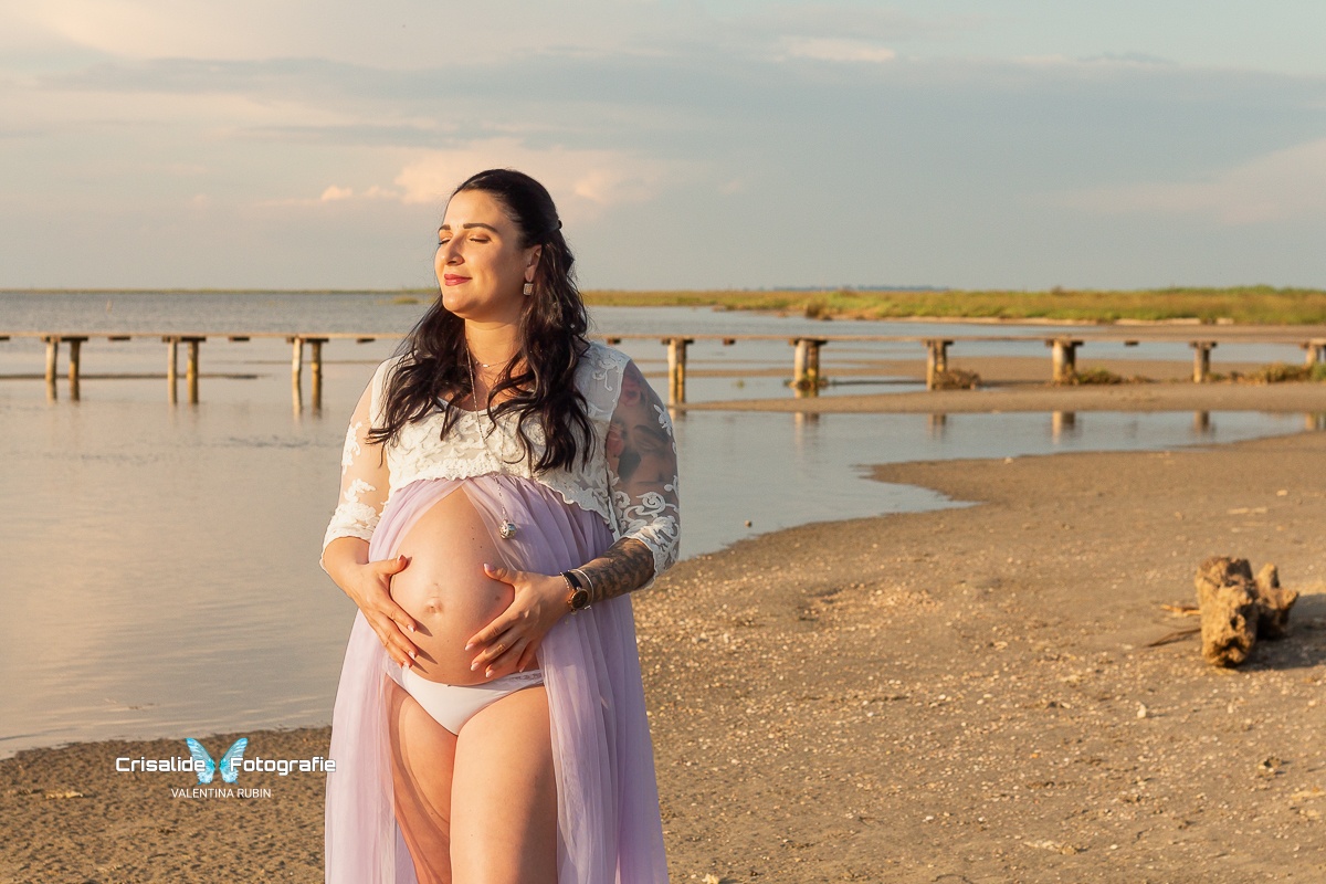 Mamma incinta con abito bianco in pizzo, maniche tre quarti, gonna in tulle lilla aperta sulla pancia. Occhi chiusi e viso rivolto al sole sorridente, mani sulla pancia, capelli neri ondulati sulle spalle. Sfondo spiaggia con tronco, mare, pontile e vegetazione, cielo azzurro con nuvole velate