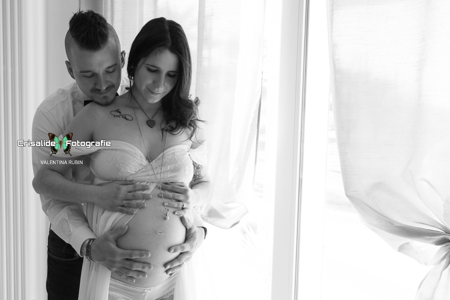 Mamma incinta in chiffon bianco e papà dietro di lei, mani sul pancione, luce entra dalla finestra a sinistra