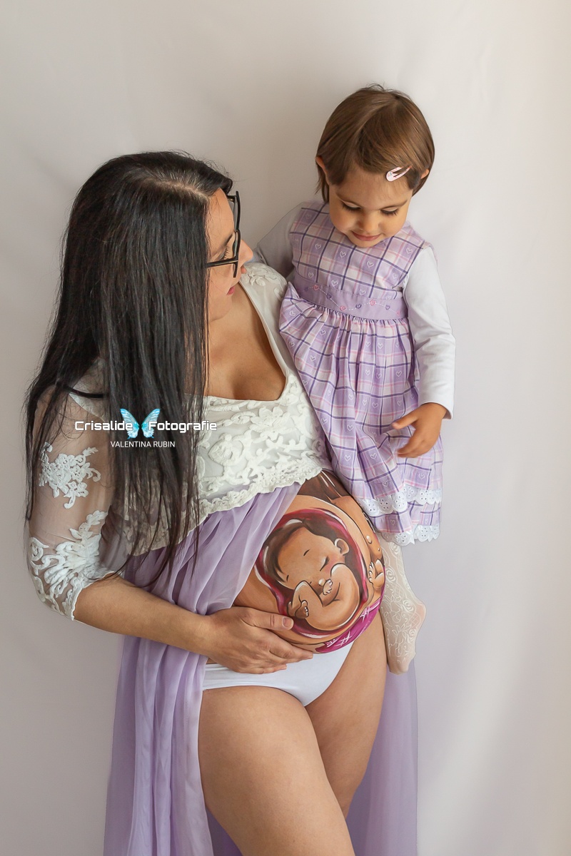 Mamma incinta con abito bianco in pizzo e gonna in tulle violetto, aperta sulla pancia, tiene in braccio la figlia di 2 anni vestita a quadretti viola e manica lunga bianca. Belly painting colorato sul pancione. La mamma con capelli neri lunghi lisci e occhiali guarda la bambina, mentre la bambina guarda il pancione. Mano della mamma sotto la pancia. Sfondo bianco, atmosfera gioiosa e familiare