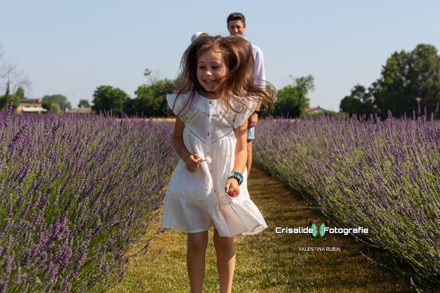 Bambina vestita di bianco che salta felice tra i filari di lavanda, con mamma e papà sullo sfondo