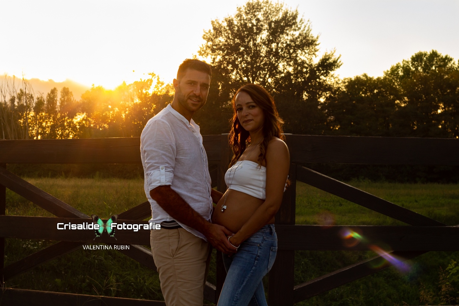Coppia che si tiene per mano e sorride all'obiettivo, lui in camicia bianca e pantaloni beige, lei con top bianco e jeans, sfondo naturale con staccionata in legno e luce del sole