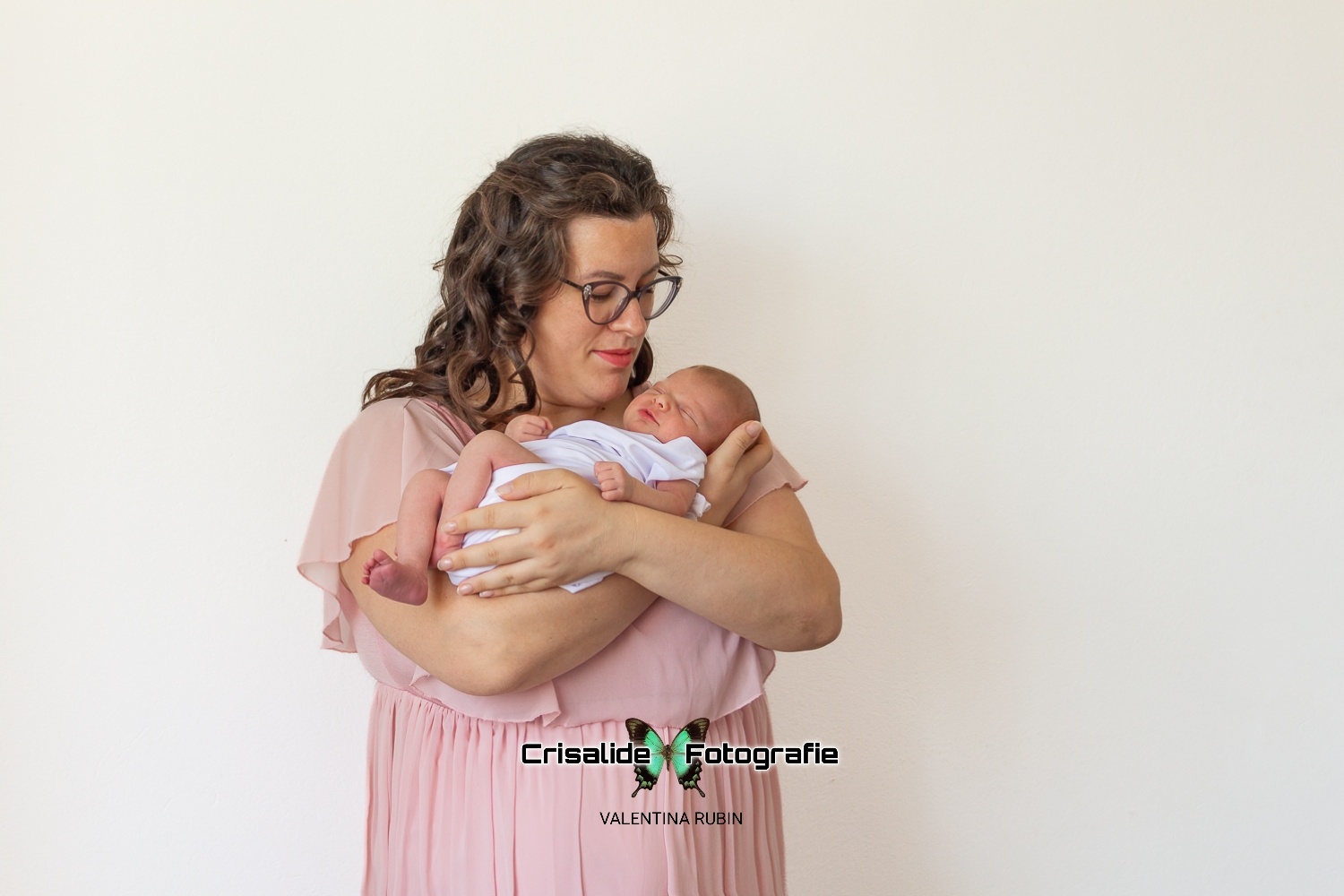 Mamma in abito di chiffon rosa che guarda sorridendo la neonata addormentata tra le sue braccia su sfondo bianco