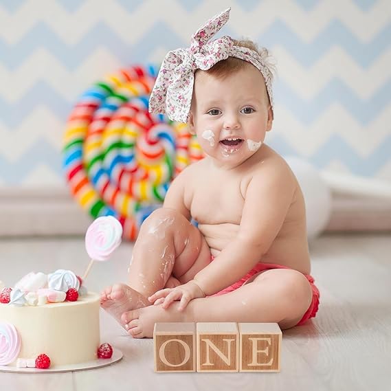 Bambina seduta a terra, con pantaloncini e petto nudo, fiocco in testa, bocca sporca da torta, di fianco una torta e cubi di legno con scritta one, sfondo colorato