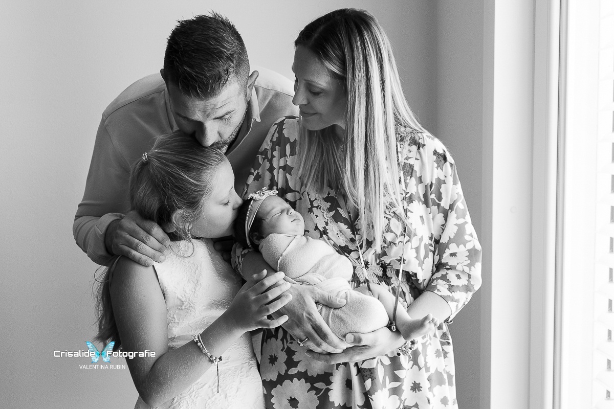 Famiglia con papà, mamma, figlia di 8 anni e neonata in un momento affettuoso in bianco e nero, illuminati dalla finestra nella loro casa