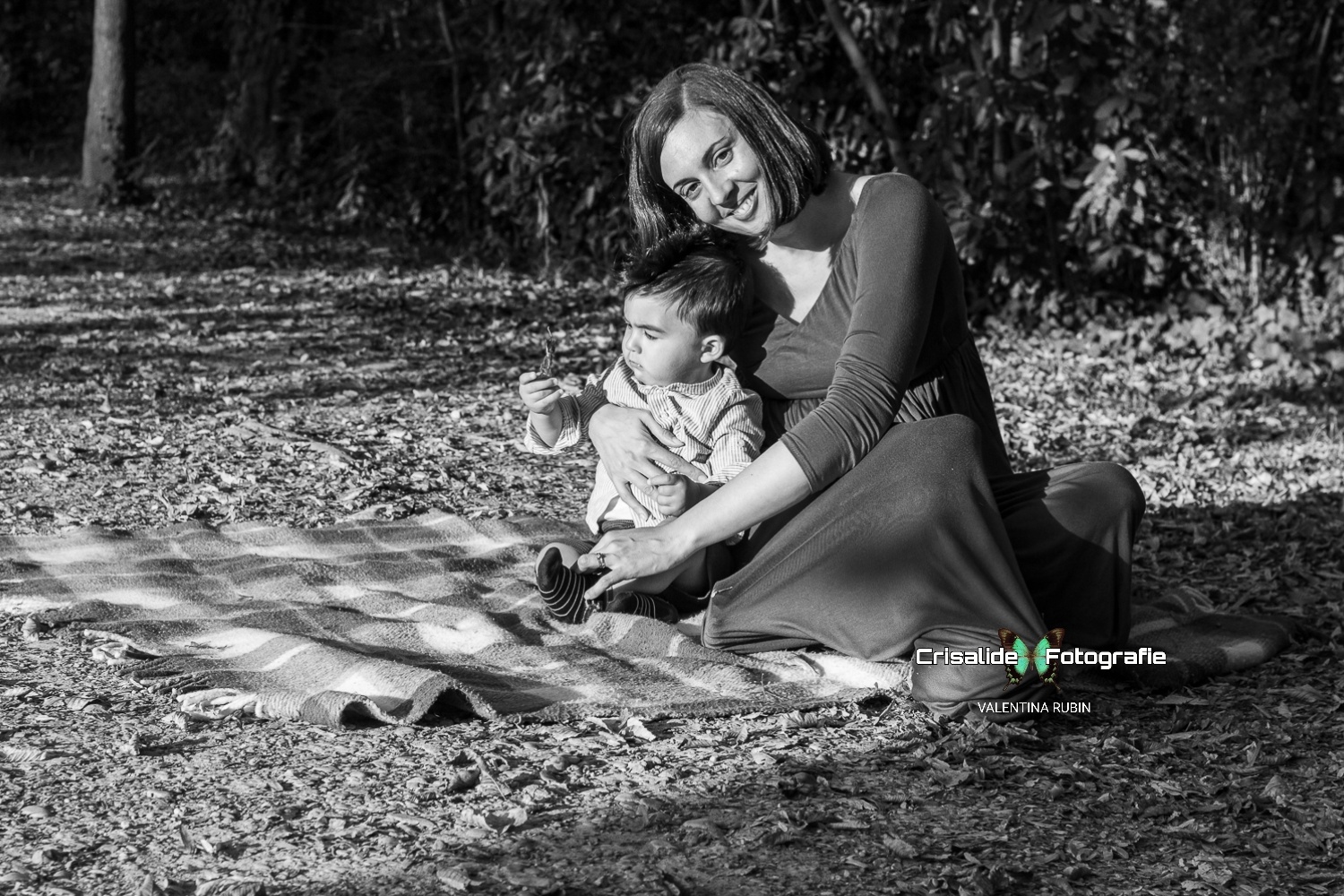 Mamma seduta su una coperta abbraccia il suo bambino mentre lui osserva una foglia secca, in un ritratto in bianco e nero con sfondo autunnale