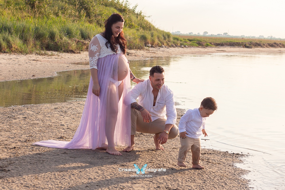 Mamma incinta in abito bianco e gonna lilla, papà accucciato in camicia bianca e pantaloni beige e bambino piccolo in coordinato con i padre, sulla spiaggia al mare. Servizio fotografico di maternità e famiglia, scatto spontaneo che cattura sorrisi, emozioni e momenti autentici di vita