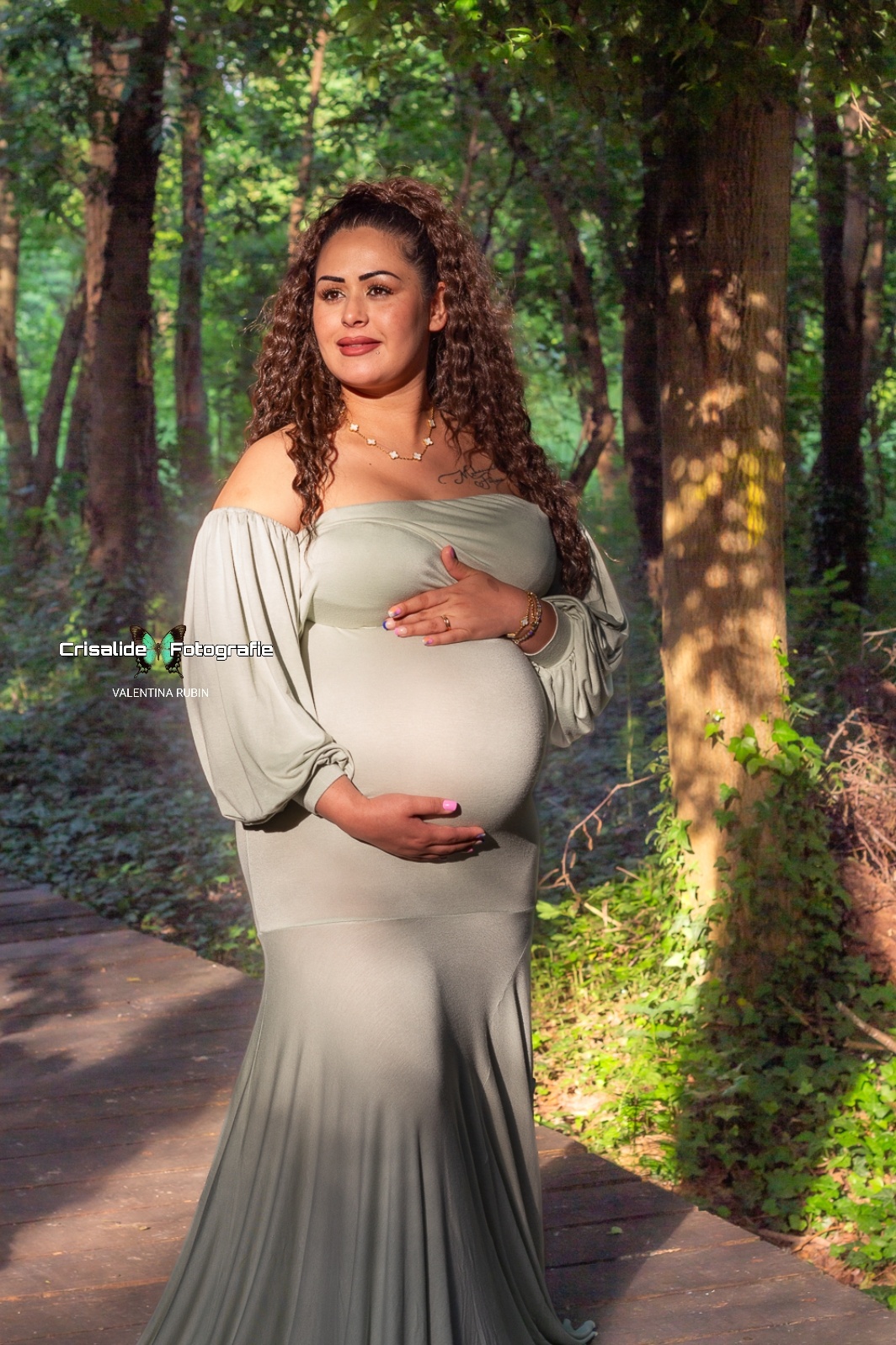 Donna incinta con abito verde chiaro a sirena, ritratta in verticale tra gli alberi. È girata di tre quarti, una mano sopra e una sotto la pancia, sorride guardando di lato. Capelli mori ondulati sulle spalle, illuminata dalla luce naturale filtrata dal verde. Scatto di maternità naturale ed emozionale