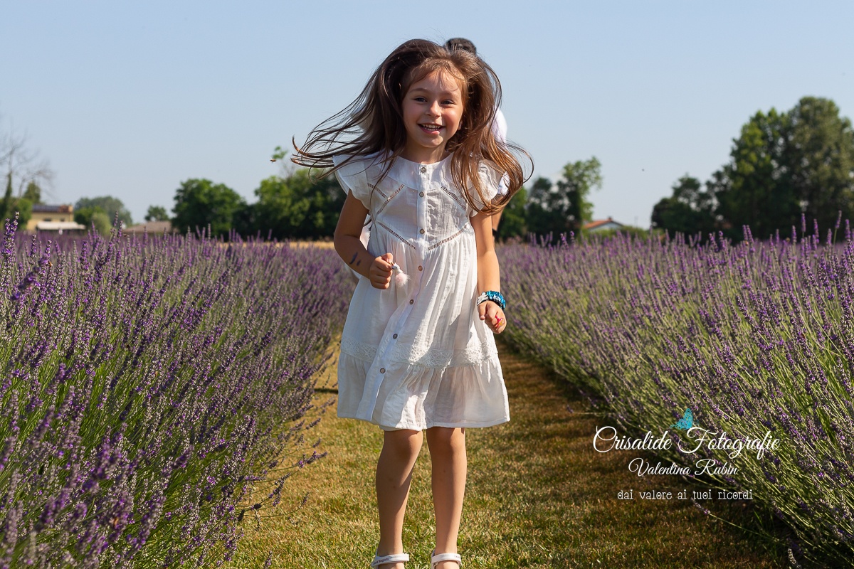 Bambina che salta sorridente tra i filari di lavanda con vestito bianco e capelli che svolazzano