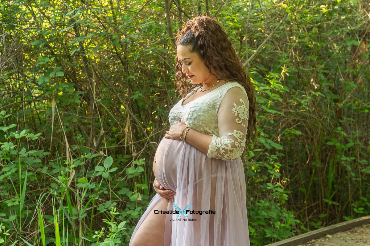 foto di maternità all'aperto, mamma incinta girata di tre quarti che guarda la pancia, una mano sopra e una mano sotto il pancione. Indossa abito in pizzo bianco con maniche tre quarti e gonna in tulle lilla leggermente aperta sulla pancia, capelli castani ondulati sulle spalle. Sfondo naturale con vegetazione verde, alberi, arbusti, e rami. Momento intimo e delicato