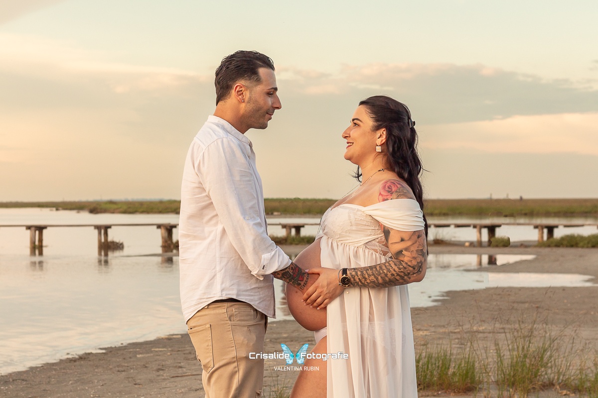 Mamma incinta di profilo in abito bianco guarda il marito sorridente, mani sulla pancia, sfondo spiaggia con mare, ciuffi d'erba e pontile