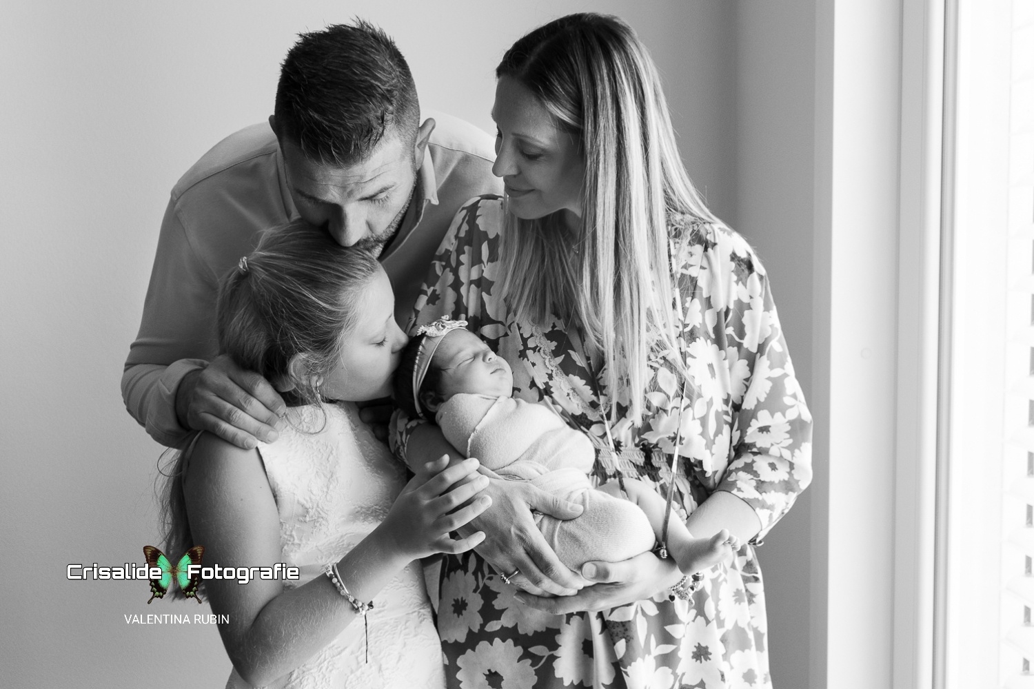 Famiglia con papà, mamma, figlia di 8 anni e neonata in un momento affettuoso in bianco e nero, illuminati dalla finestra nella loro casa