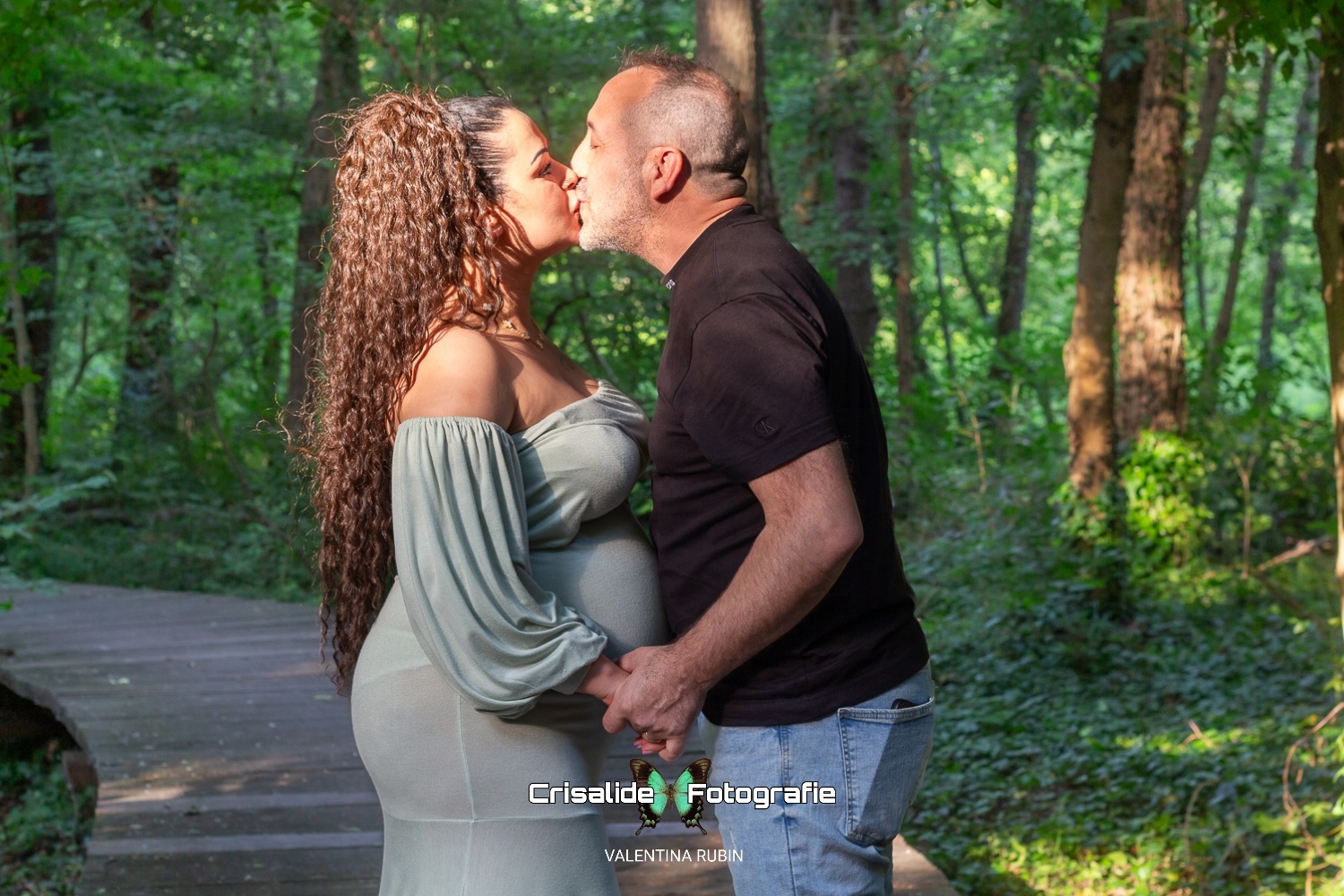 Coppia che si bacia tenendosi per mano lungo un sentiero nel bosco, lei con abito verde chiaro e capelli ondulati, lui con maglia nera e jeans, circondati da alberi