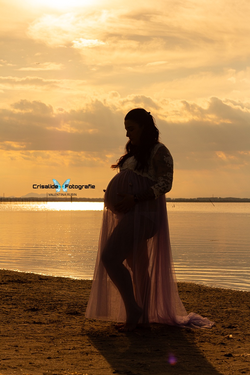 Foto in controluce, soggetto in penombra. Silhouette di mamma incinta, girata di tre quarti sulla spiaggia, mani sotto la pancia. Abito bianco in pizzo, manica tre quarti, gonna in tulle lilla. Figura intera con piedi sulla sabbia. Mare sullo sfondo con riflessi dorati del sole e nuvole luminose. Atmosfera magica, luminosa e naturale al tramonto
