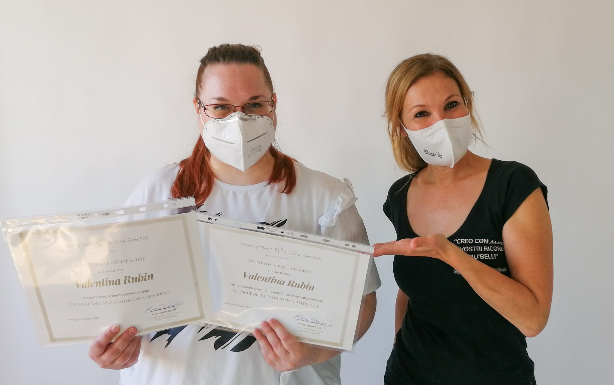 Valentina Rubin insieme alla fotografa Elisa Spreafichi durante il mentoring di fotografia di maternità e neonati, entrambe con mascherina, fatto in periodo covid, e sorridenti con gli occhi; Valentina tiene in mano gli attestati mentre Elisa li indica con la mano, su sfondo bianco
