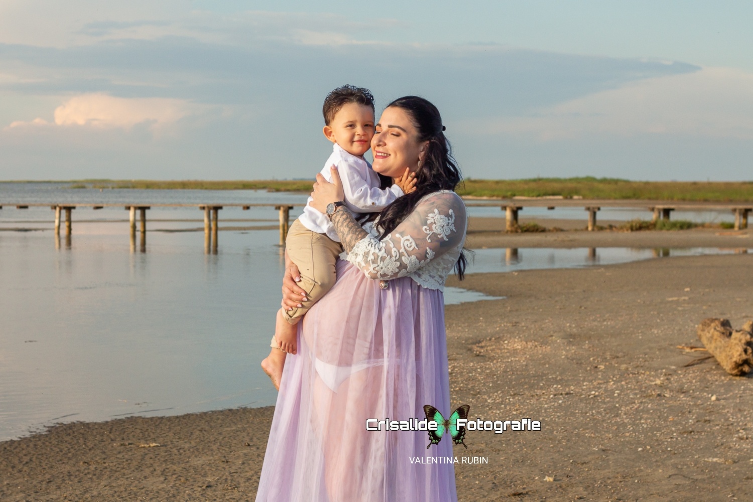 Mamma incinta in abito bianco di pizzo tiene in braccio il bambino vestito di bianco e beige, sorridono, sfondo spiaggia, tronco, mare e pontile