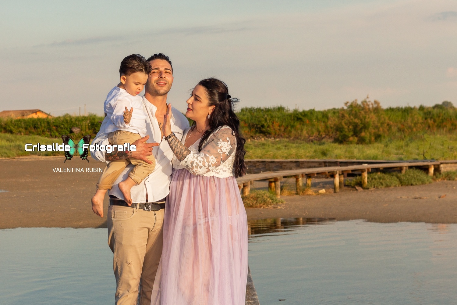 Mamma incinta sulla destra con abito bianco in pizzo e gonna in tulle lilla, mano alzata per dare il cinque al figlio. Papà al centro con camicia bianca e pantaloni beige, in braccio il bambino di circa 1 anno e mezzo con abbigliamento coordinato con papà. Papà sorridente con occhi chiusi illuminato dal sole. Bambino con manina alzata verso la mamma. La famiglia è sul pontile, con mare ai lati, spiaggia e vegetazione sullo sfondo e cielo azzurro