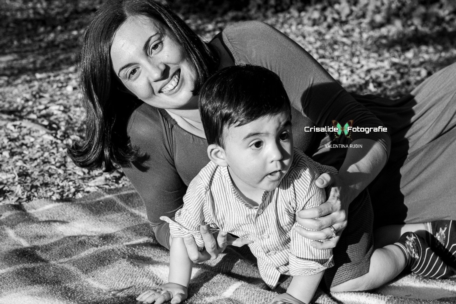 Mamma sdraiata su una coperta sorride all'obiettivo mentre tiene tra le braccia il suo bambino che, a gattoni, esplora l'ambiente intorno, in uno scatto in bianco e nero