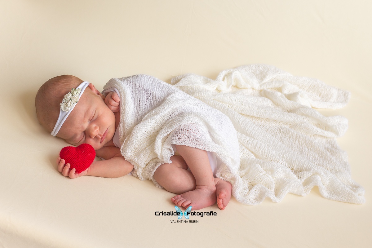 Neonata che dorme di lato con fascetta floreale, cuore rosso tra le mani, avvolta in tessuto bianco su sfondo beige