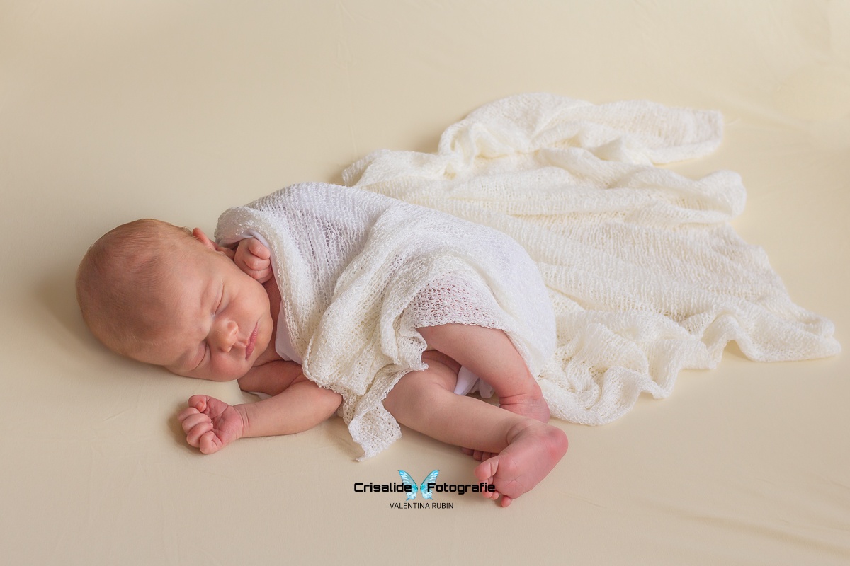 Neonata che dorme di lato avvolta in uno scialle bianco ricamato su sfondo beige