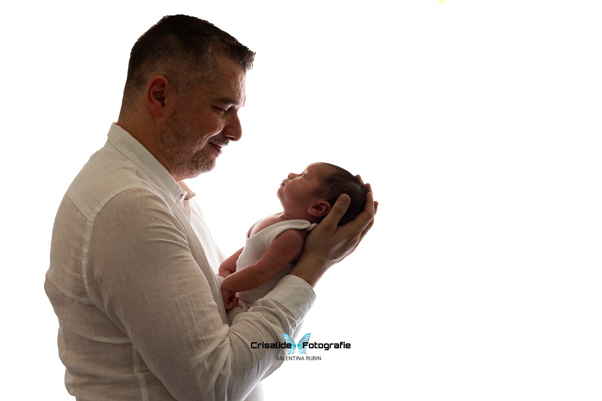 Papà sorride guardando il neonato vestito con body bianco, testina sorretta dalle mani del papà, sfondo bianco