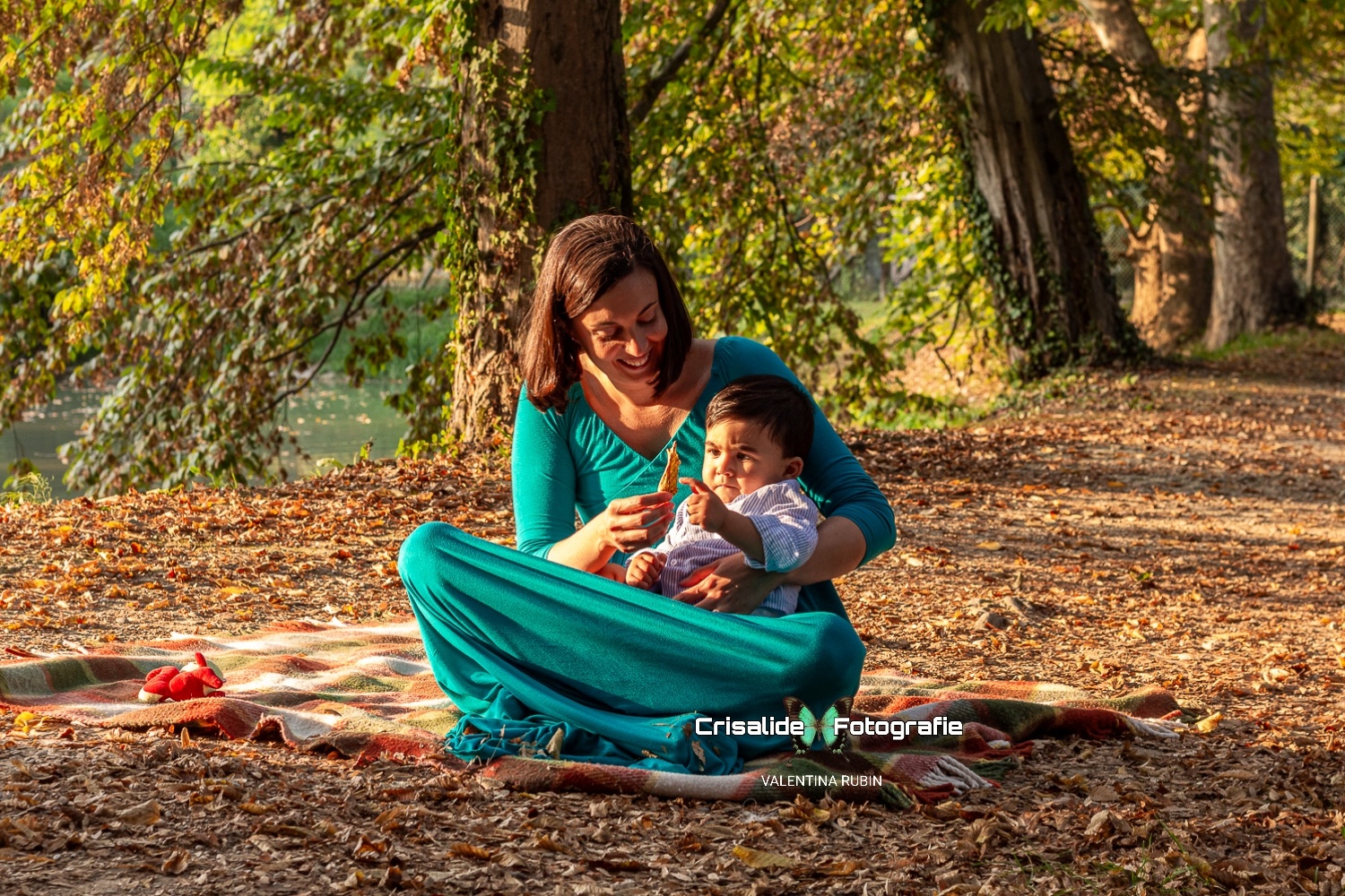 Mamma seduta su una coperta con il suo bambino in braccio, che osserva curioso una foglia secca, immersi in un paesaggio autunnale