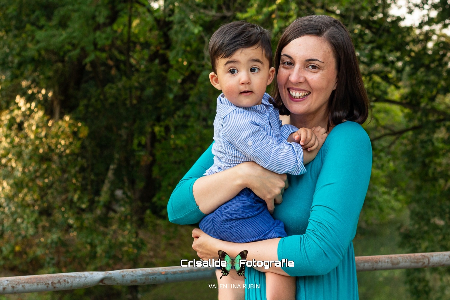 Mamma con vestito lungo verde, sorride verso l'obiettivo, tiene in braccio il bambino con pantaloncini blu e camicia a righe azzurre, lui guarda di lato. Sfondo alberi