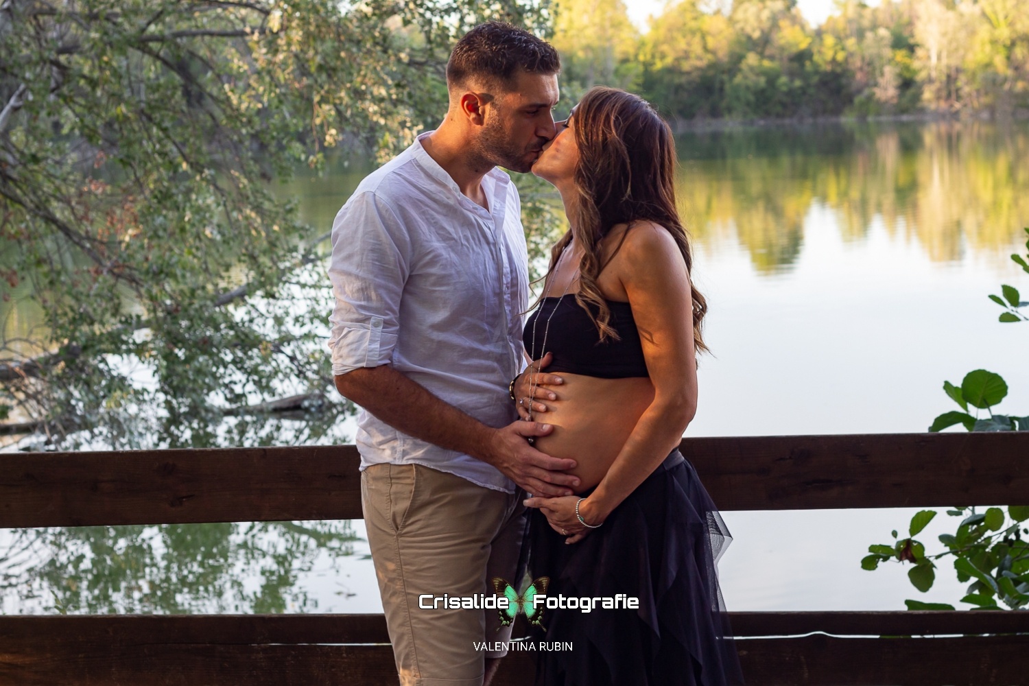Coppia che si bacia, lui in camicia bianca e pantaloni beige, lei con top nero e gonna in tulle, sfondo naturale con staccionata in legno, laghetto e alberi