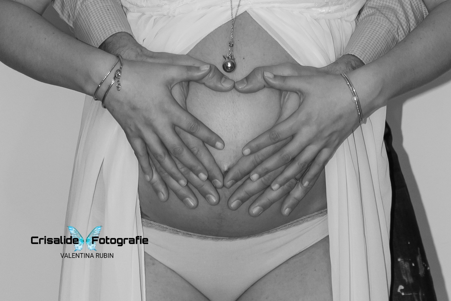 Primo piano del pancione di mamma, mani del papà e della mamma formano un cuore a chiudersi sull'ombelico, ciondolo appena sopra al cuore, braccialetti ai polsi della mamma