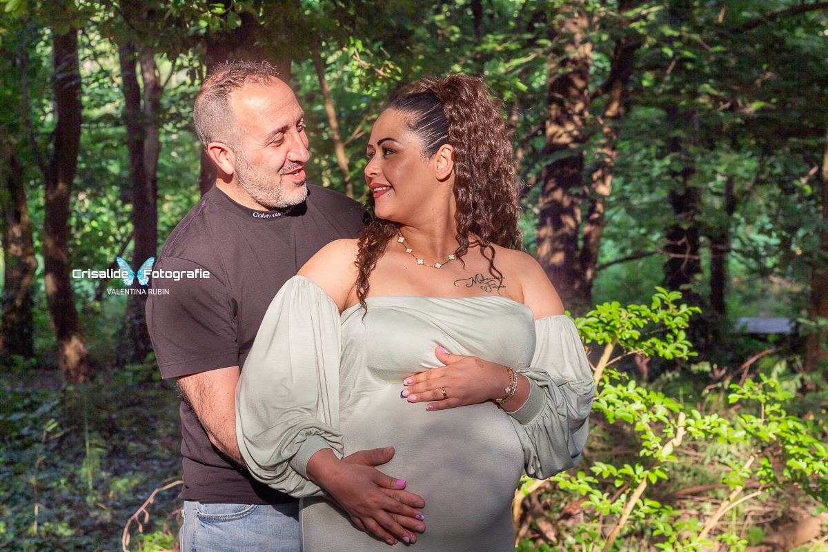 Mamma incinta vestita in abito verde chiaro a sirena, maniche lunghe e spalle scoperte, viso girato verso sinistra sorridente che guarda il marito. Una mano sopra e una sotto la pancia, mano del marito sopra la sua. Marito dietro di lei a sinistra, maglia nera e jeans, sorriso rivolto a lei. Illuminati da luce solare filtrata dagli alberi, sfondo naturale con alberi e vegetazione