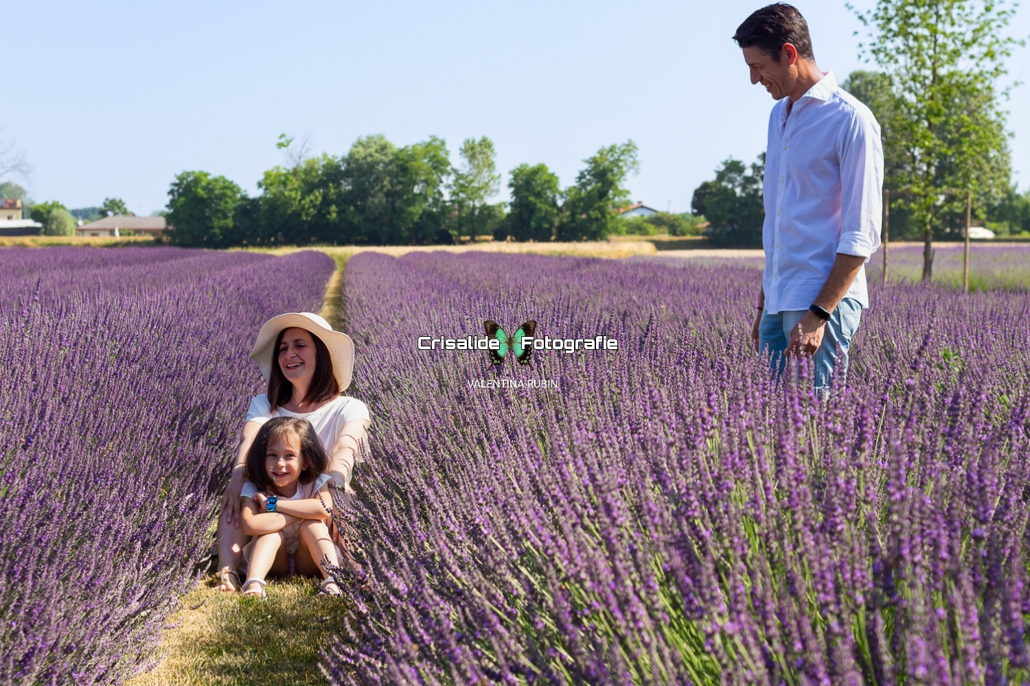 Mamma e figlia sedute tra i filari di lavanda, vestite di bianco, mentre il papà è in piedi accanto a loro sorridente, guardandole con affetto
