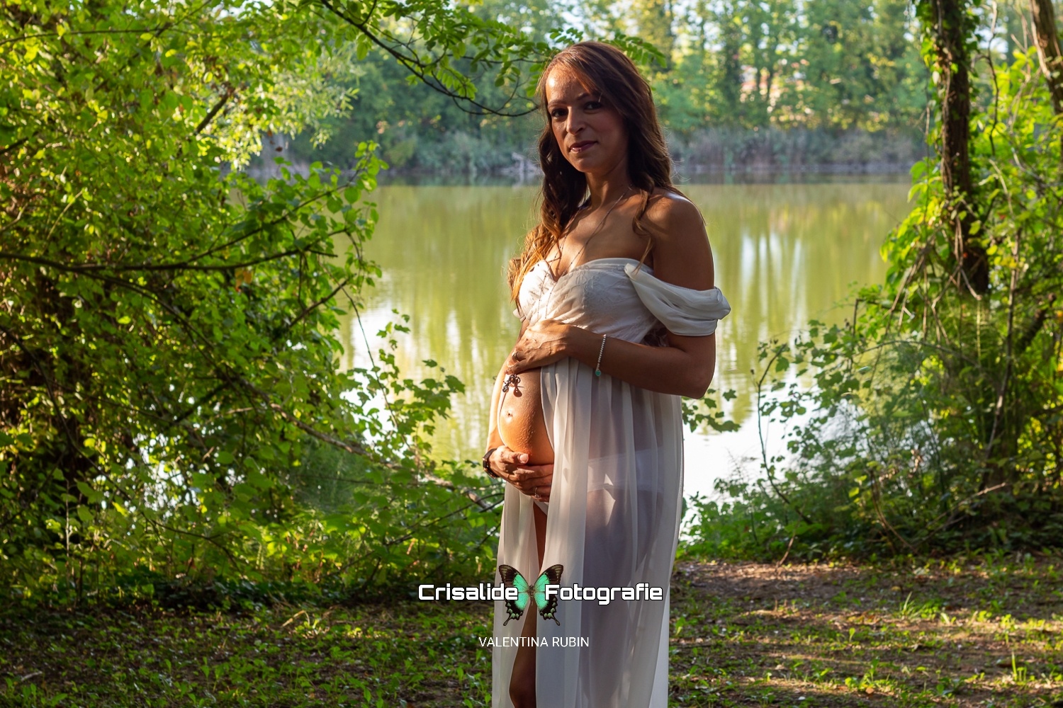 Mamma incinta girata di tre quarti con abito bianco in chiffon, spalle scoperte e apertura sulla pancia, sguardo sorridente verso la camera. Capelli castani ondulati sulle spalle, ciondolo sulla pancia. Illuminata da luce solare e ombreggiata da alberi. Sfondo naturale con scorcio di laghetto