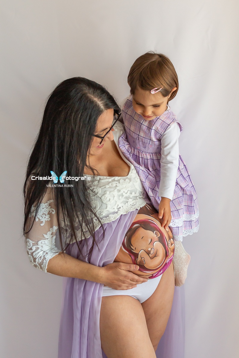 Mamma incinta con belly painting che tiene in braccio la figlia di 2 anni; entrambe guardano sorridenti la pancia, su sfondo bianco