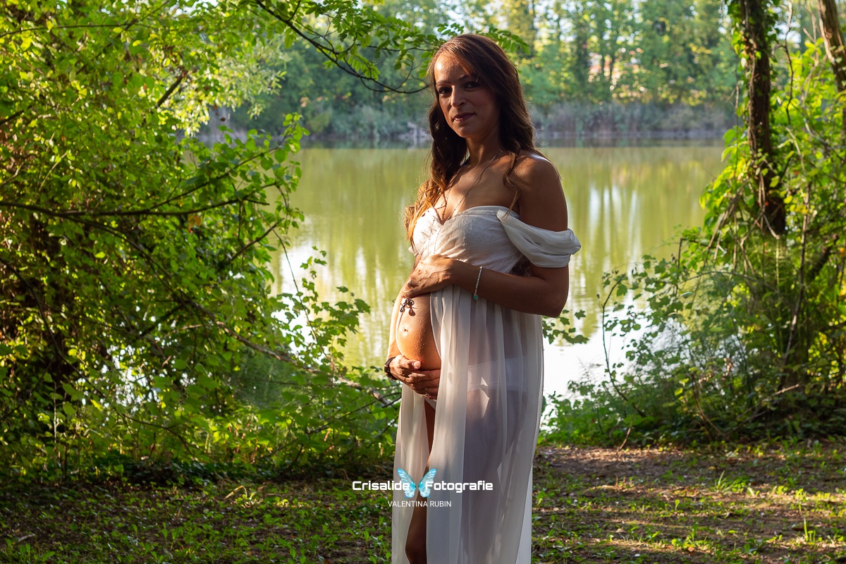 Mamma incinta girata di tre quarti con abito bianco in chiffon, spalle scoperte e apertura sulla pancia, sguardo sorridente verso la camera. Capelli castani ondulati sulle spalle, ciondolo sulla pancia. Illuminata da luce solare e ombreggiata da alberi. Sfondo naturale con scorcio di laghetto